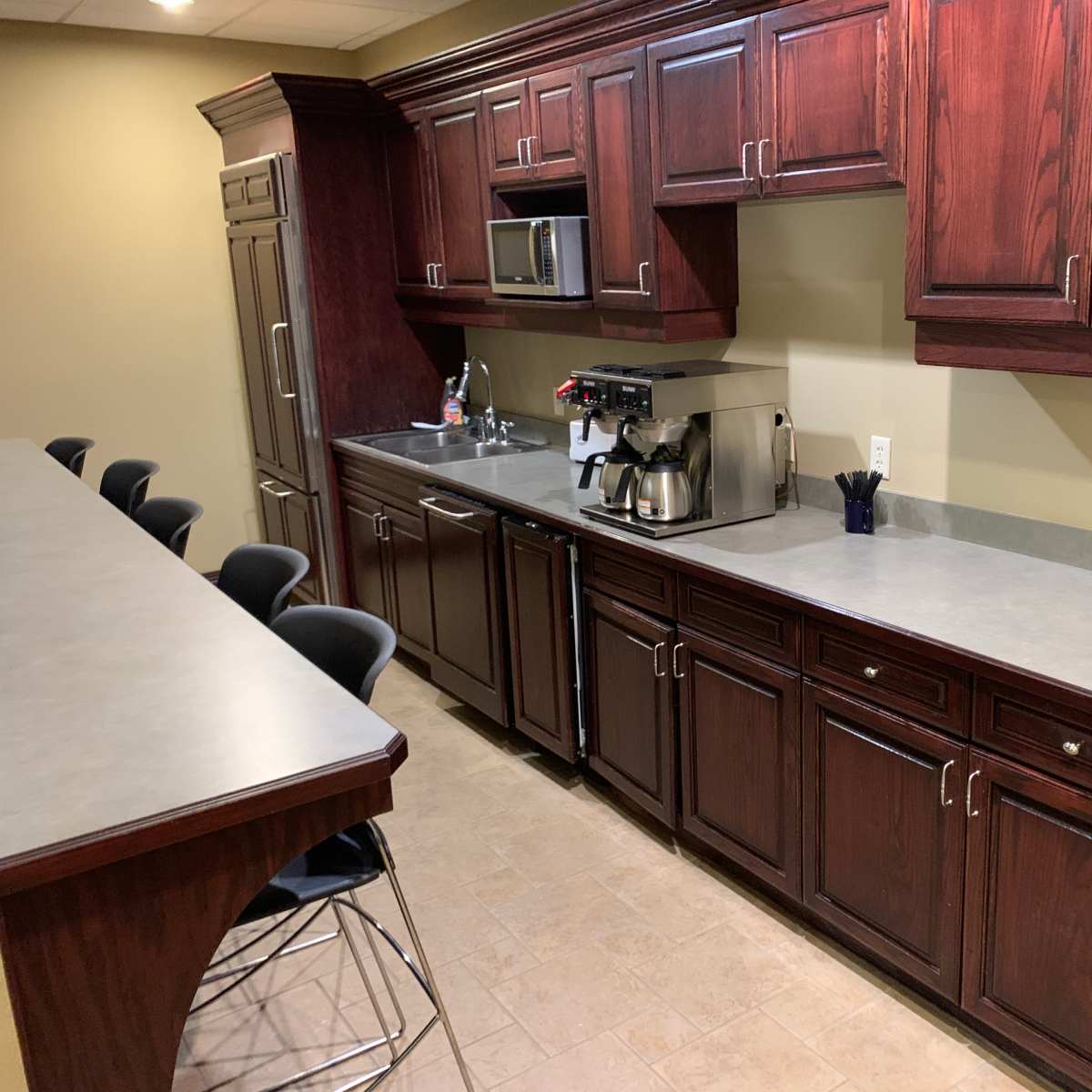 A modern kitchen with dark wooden cabinets, a countertop, and a coffee machine, positioned next to a bar with high stools.