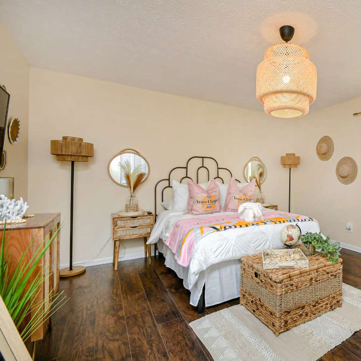 A well-decorated bedroom featuring a bed with colorful bedding, two bedside tables, a woven bench, and a large window allowing natural light to enter.