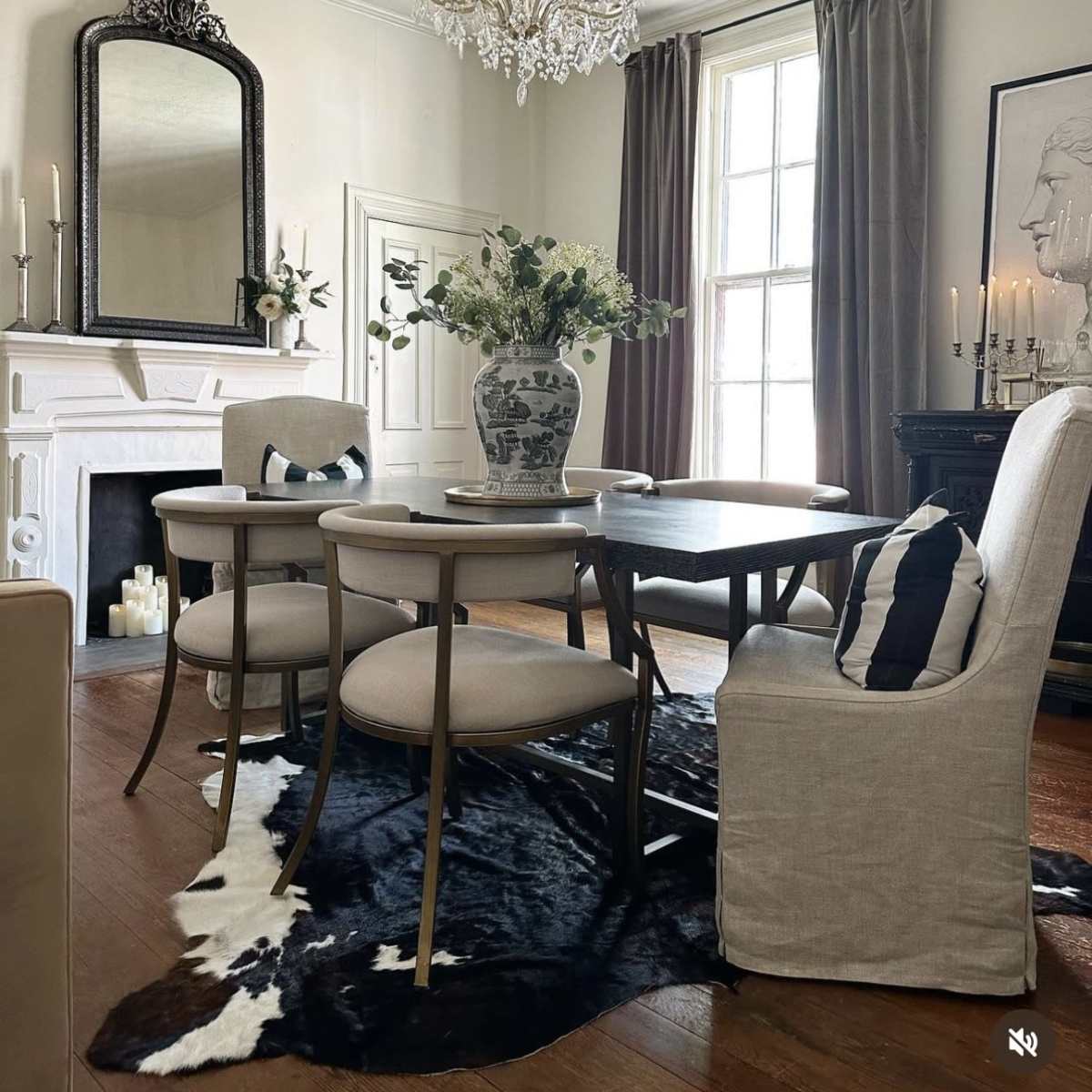A dining room features a wooden table surrounded by upholstered chairs, a large vase with flowers on the table, and a chandelier above, with a decorative fireplace and art on the walls.