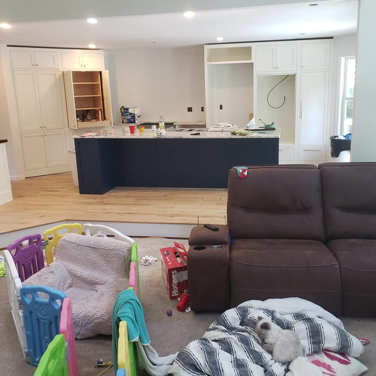 A messy living room with a brown couch, scattered toys, a dog on a blanket, and an unfinished kitchen area in the background.