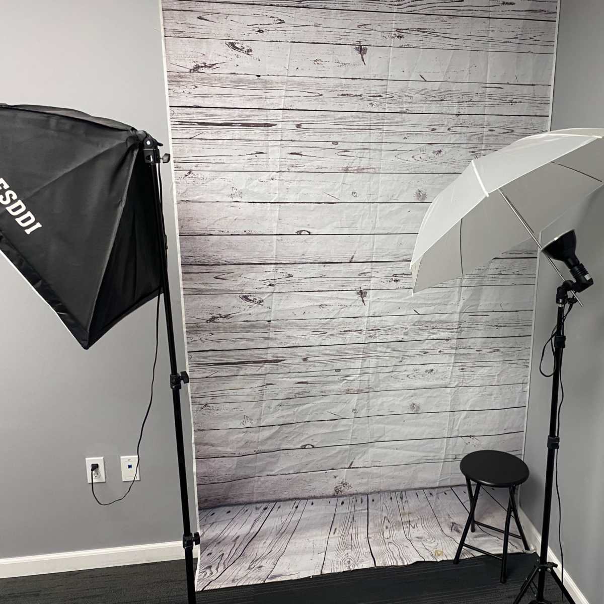 A photography setup featuring a backdrop of wooden planks, a lighting umbrella, and a black stool.