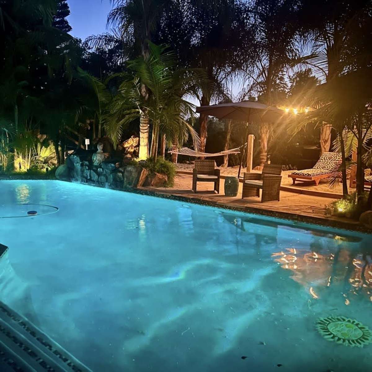 A lit swimming pool surrounded by palm trees and lounge chairs in a serene outdoor setting at dusk.