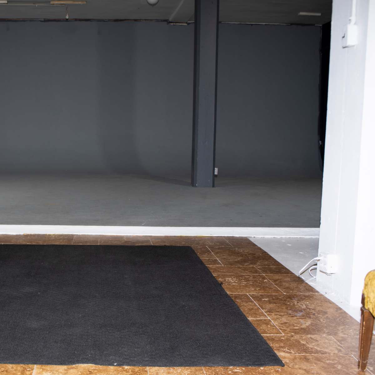 An empty room with a gray background, a black rug on a tiled floor, and a wooden cabinet with audio equipment on the left.