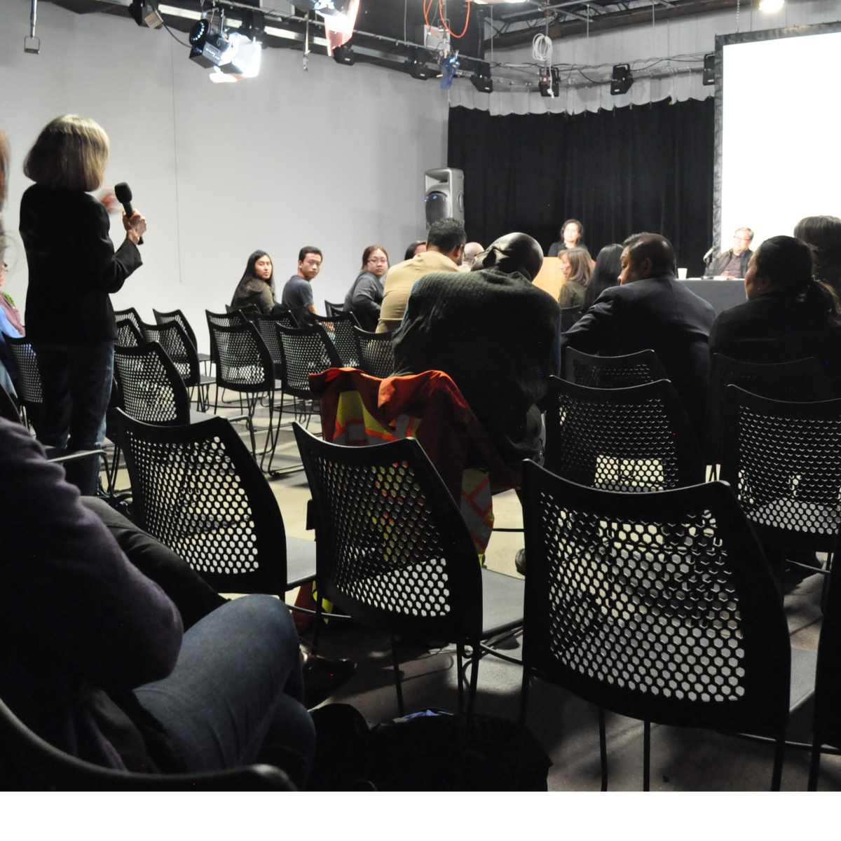 A woman holds a microphone while standing in front of a seated audience during a panel discussion in a dimly lit room.