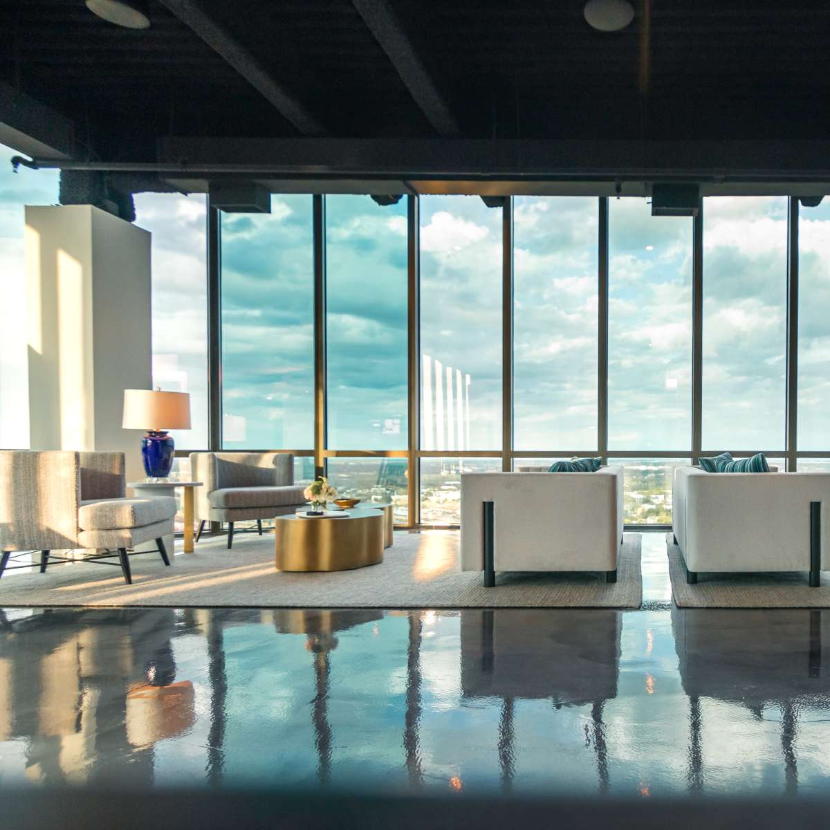 A modern lounge area with large windows, two white sofas, and decorative tables against a reflective floor.