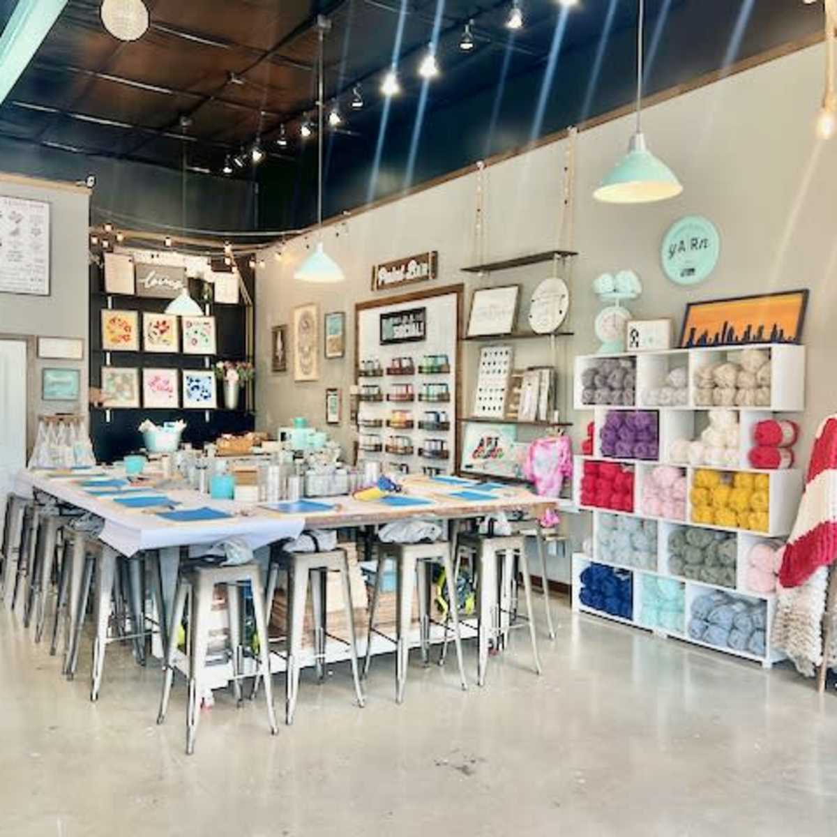 An art studio with a long table set up for crafting, surrounded by shelves filled with colorful yarn and various art supplies.