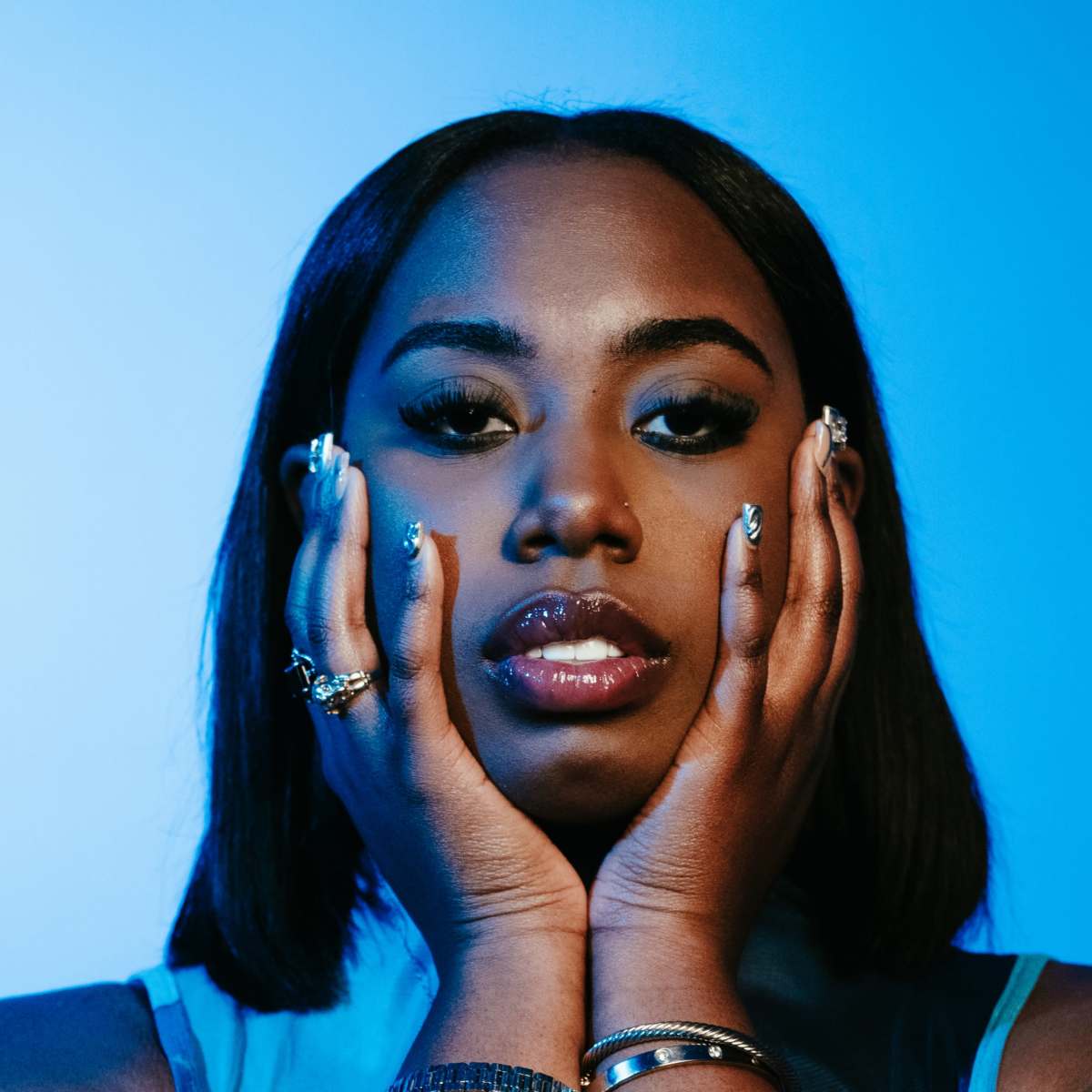 A woman with long, straight hair poses with her hands on her face against a blue background.