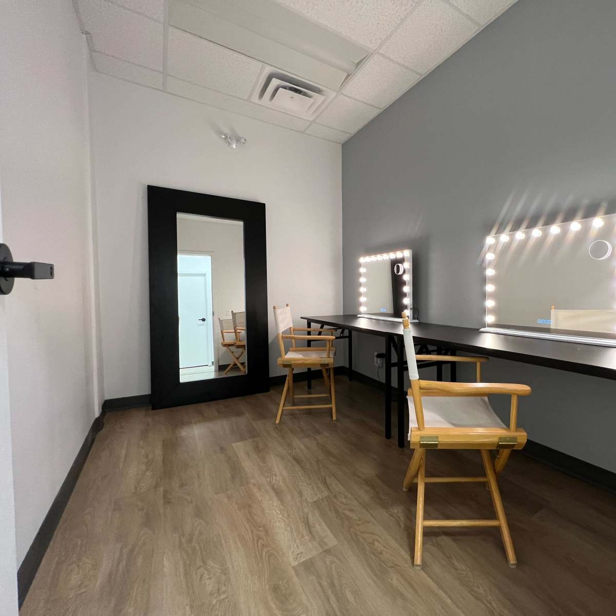 A simple makeup room featuring two lighted mirrors, two chairs, and a full-length mirror against a gray wall.