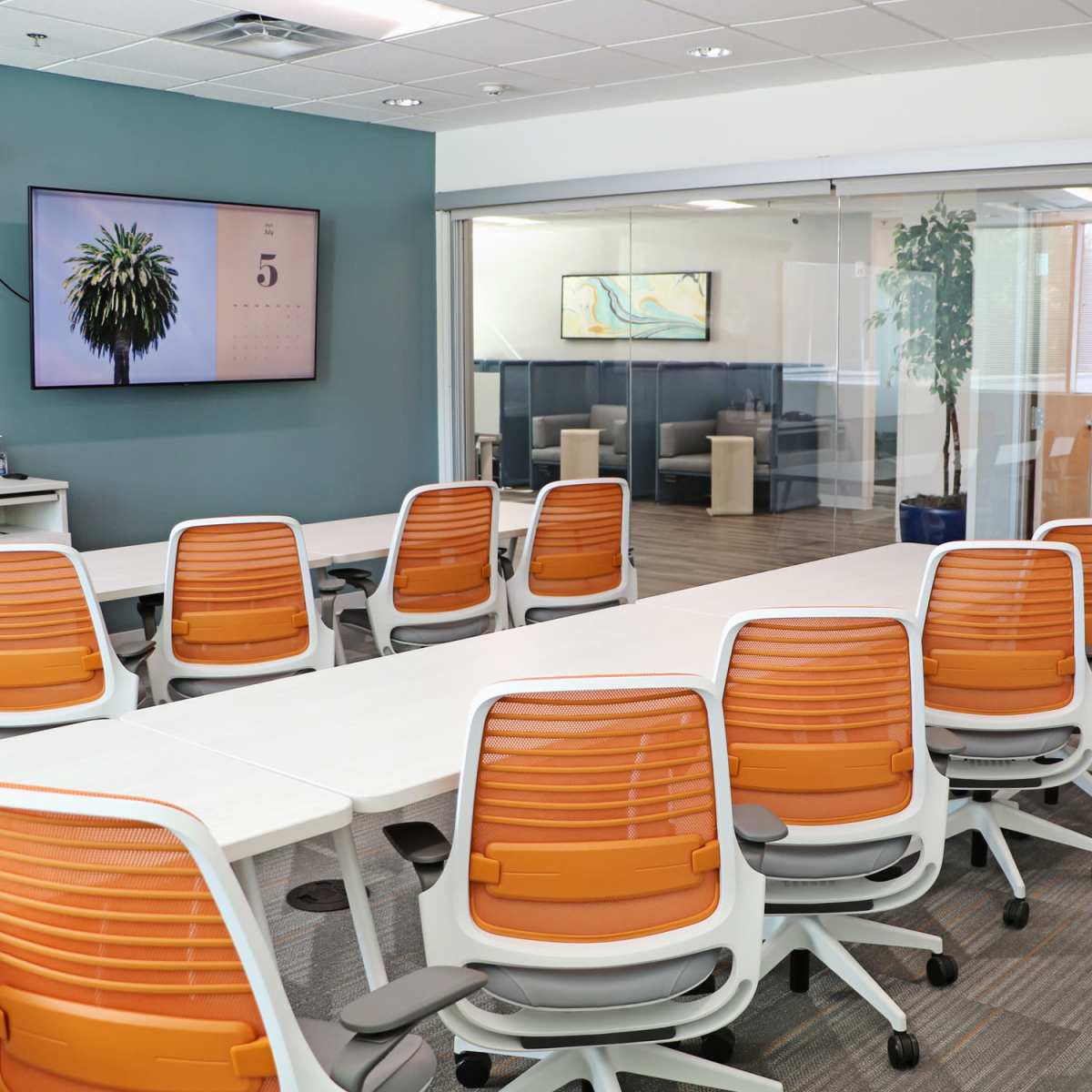 A modern conference room with white tables and orange office chairs, featuring glass walls and a large wall-mounted monitor displaying an image of a plant.