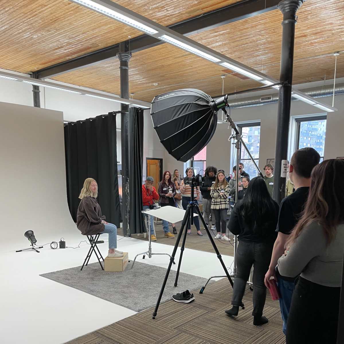 A group of people observes a photoshoot setup featuring a seated model in front of a lighting umbrella against a neutral backdrop.