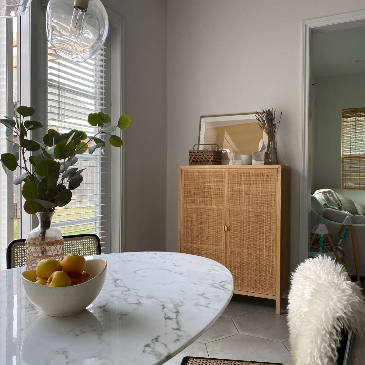 A round marble dining table is set with a bowl of fruit and surrounded by chairs, with a wooden cabinet and decorative items visible in the background.
