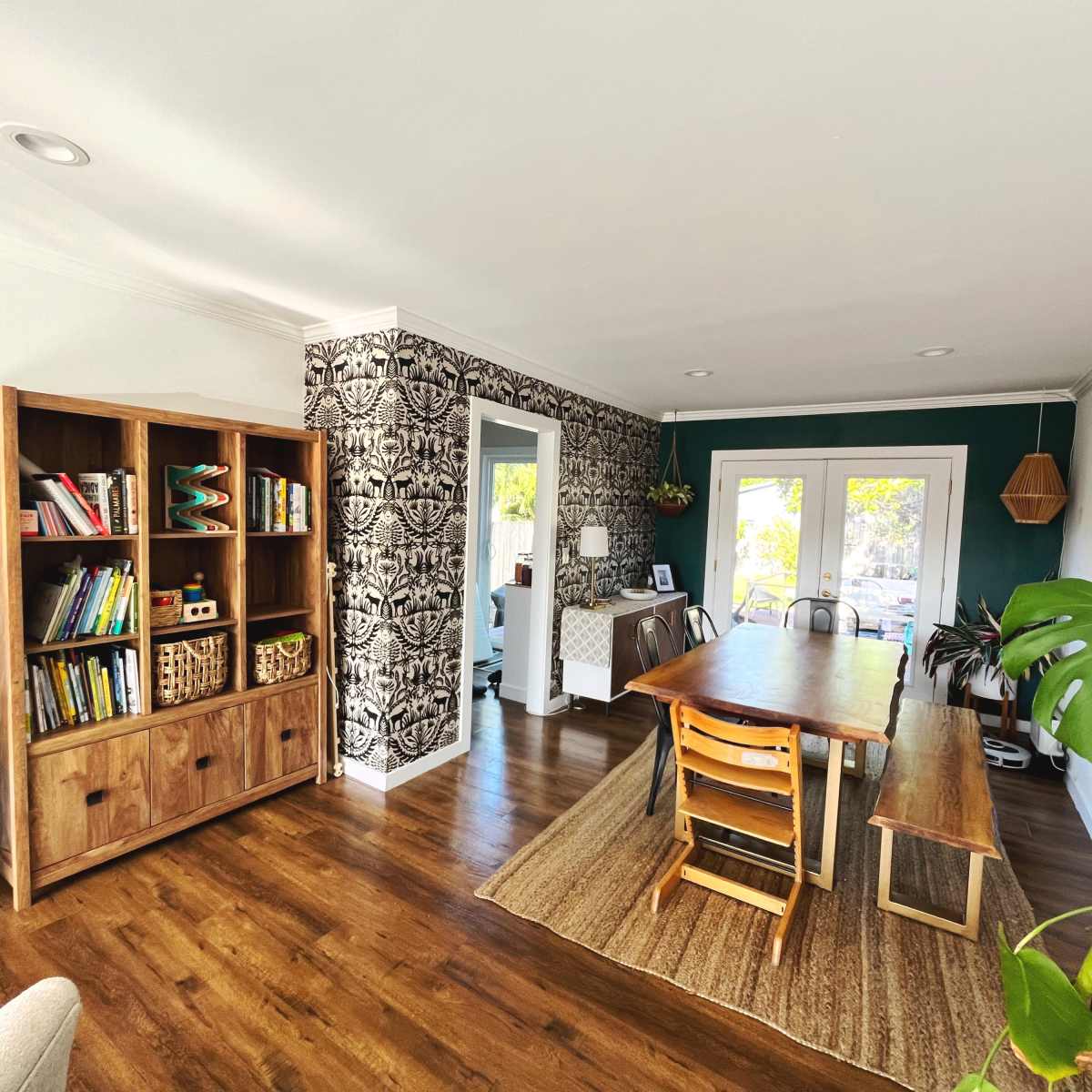 A modern living space featuring a wooden bookshelf filled with books, a dining table with chairs, and patterned wallpaper in a corner.