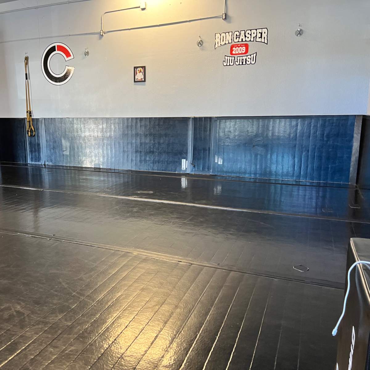 A spacious jiu-jitsu training mat area with black flooring and a wall displaying an American flag and gym branding.