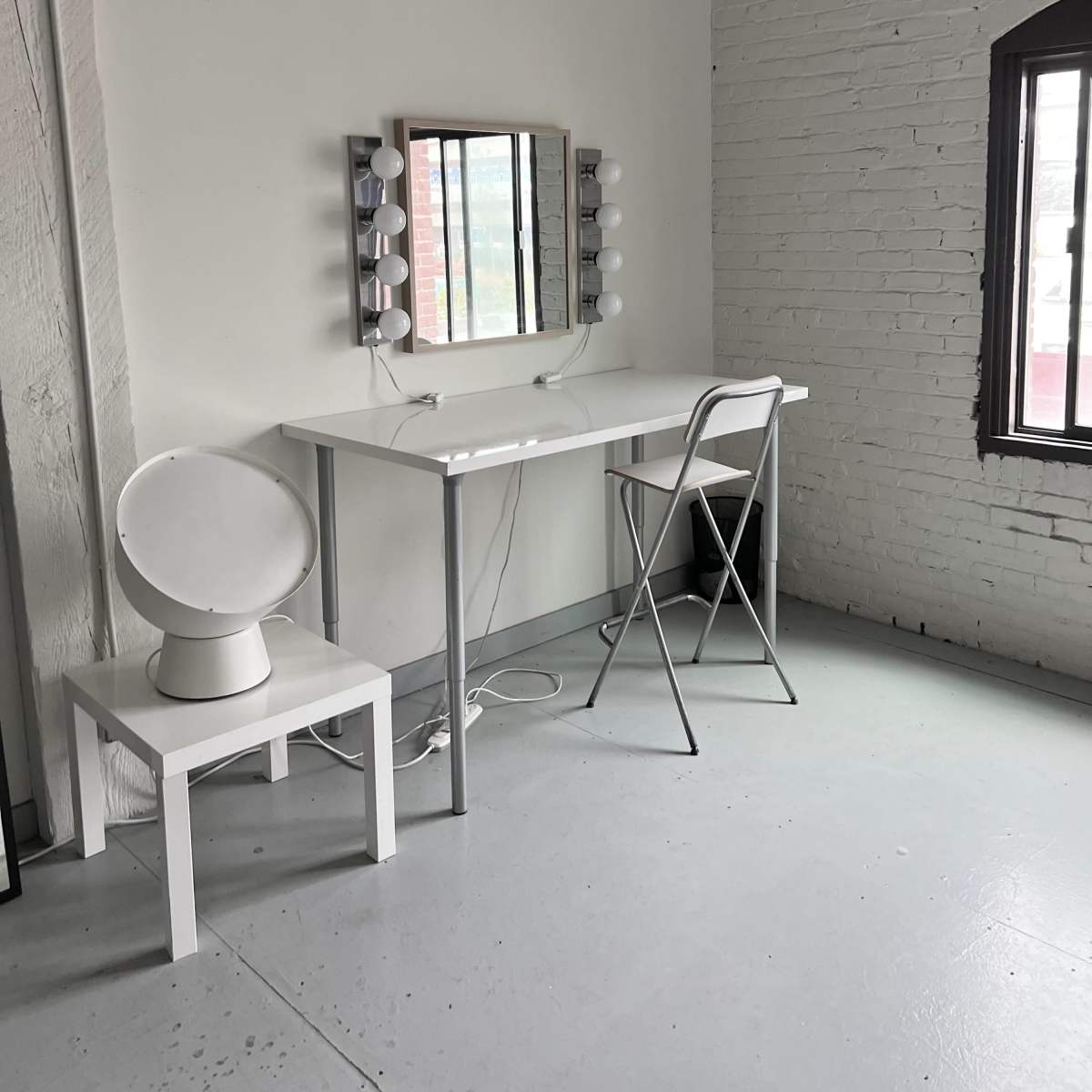 A minimalist makeup vanity with a mirror and lights sits against a white brick wall beside a chair and circular stool in a well-lit room.