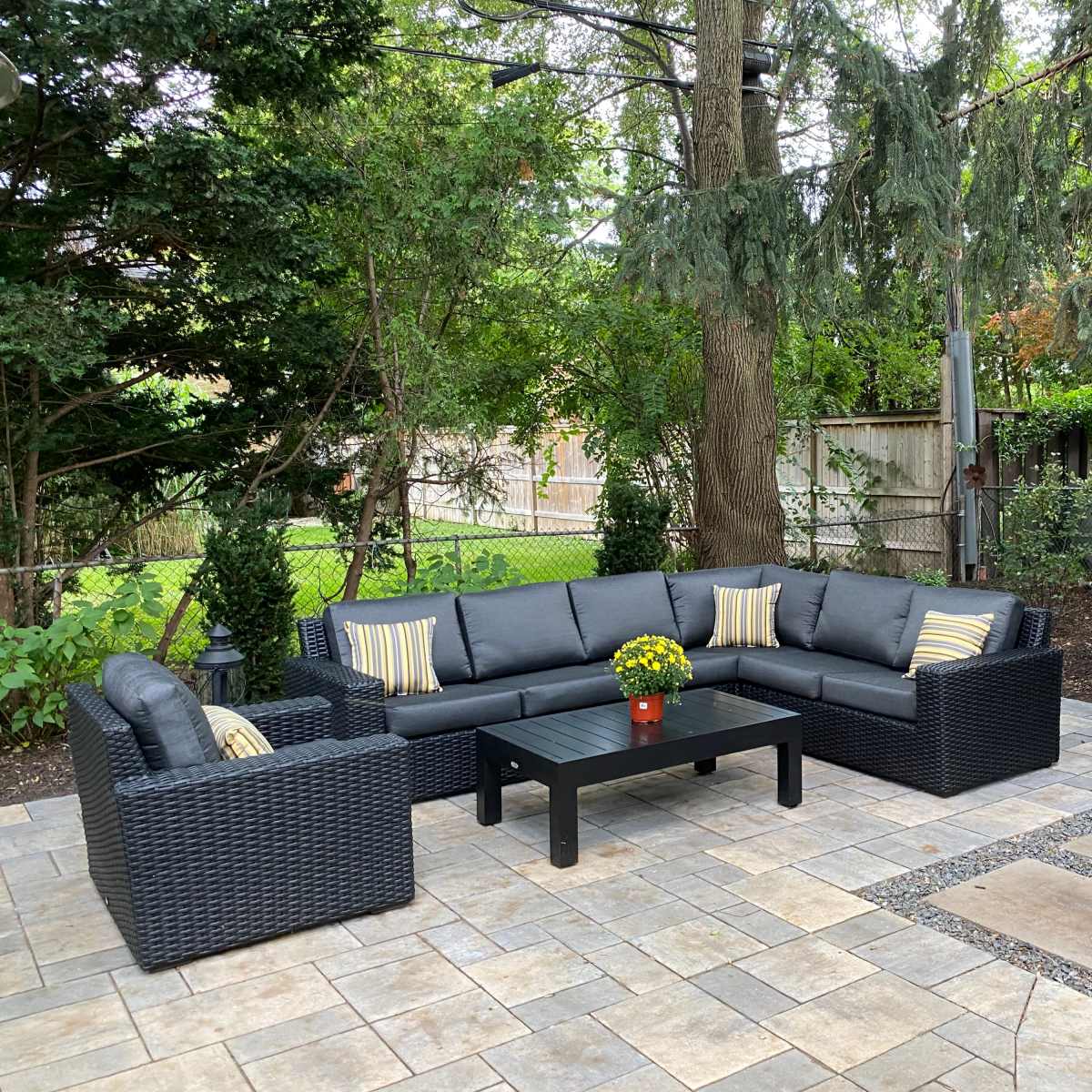 A patio area features a black wicker seating set with cushions, a black coffee table, and a flower pot, surrounded by greenery and wooden structures.