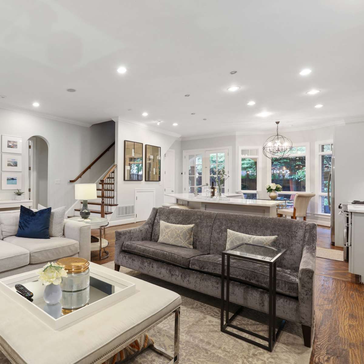 A spacious living room with two sofas, a coffee table in the center, and a staircase leading to an upper level, all illuminated by recessed lighting.