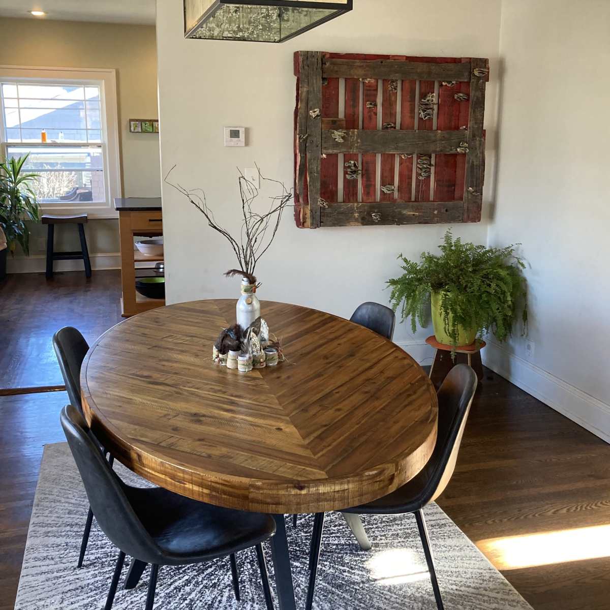 A round wooden table is set with a vase and decorations, surrounded by black chairs, with a red and wooden wall art nearby and a plant in a pot.