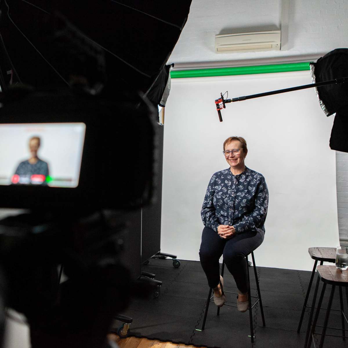 A person sits on a stool in front of a plain backdrop while being filmed by a camera with studio lighting.