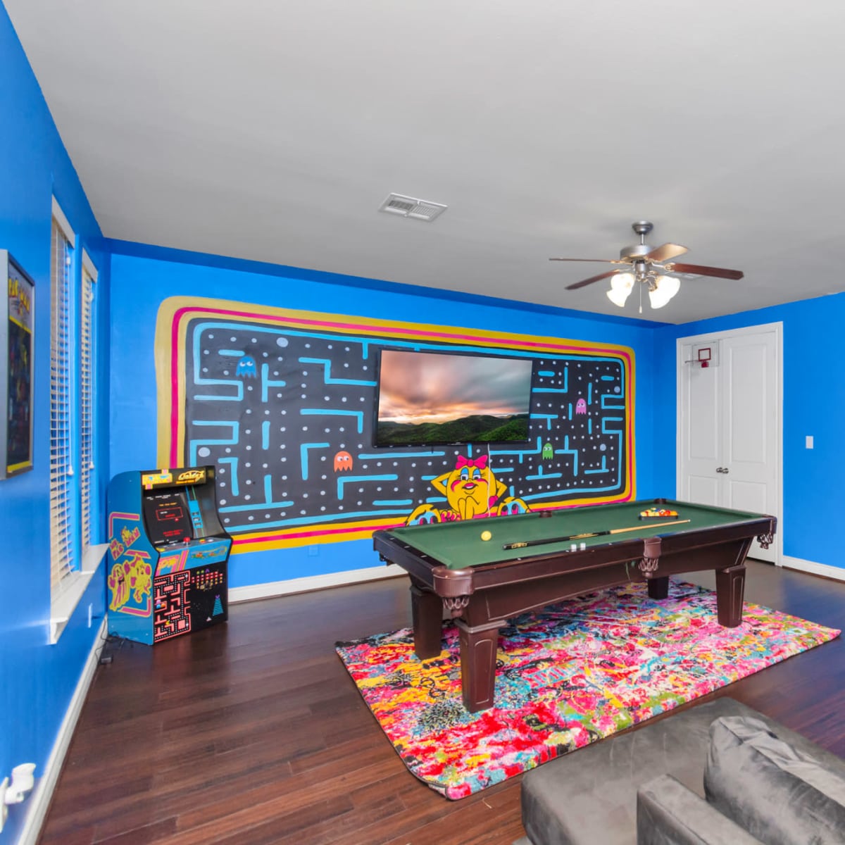 The room features a pool table in front of a colorful wall with a Pac-Man mural and an entertainment setup.