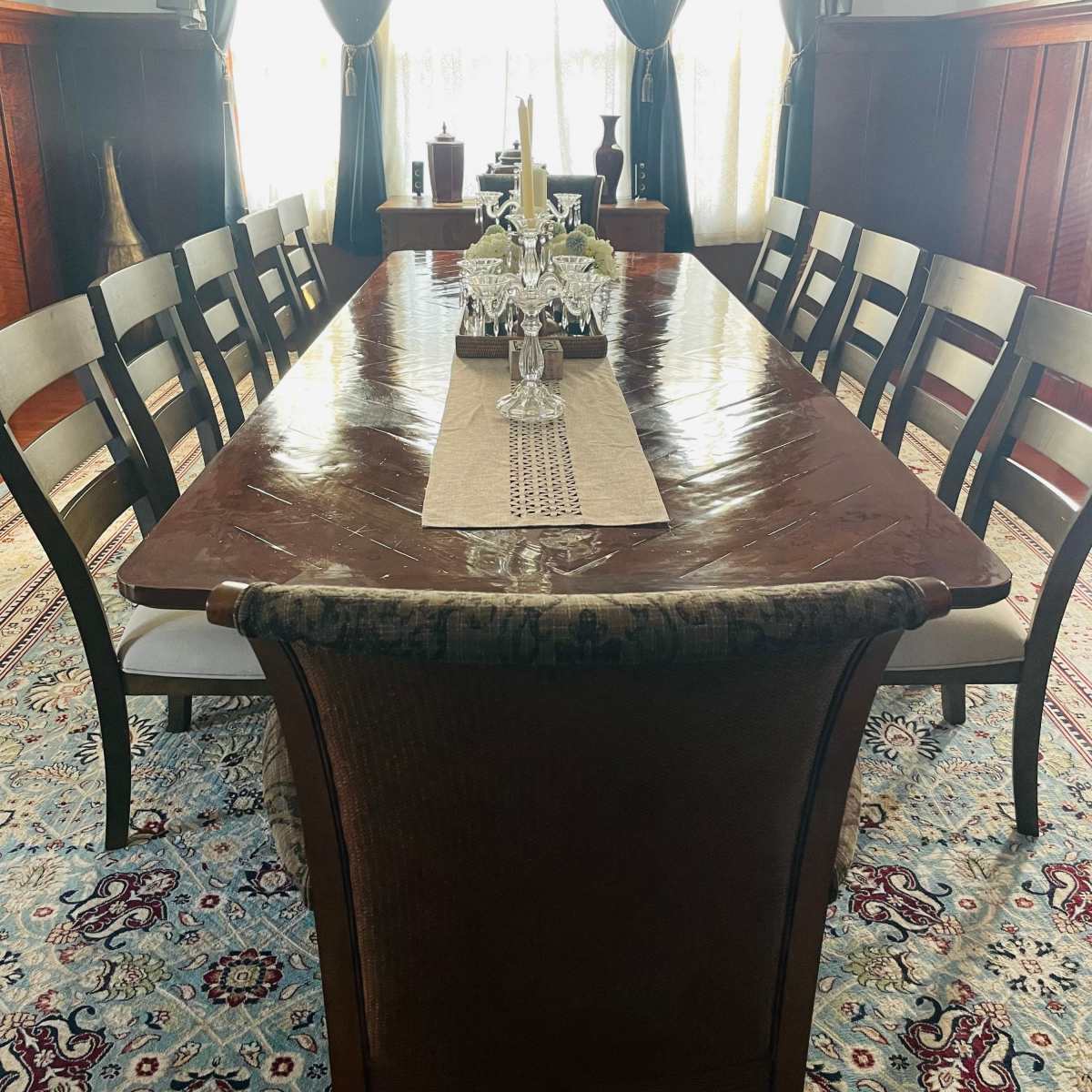 A long wooden dining table is set in a well-lit room with a chandelier and decorative curtains, surrounded by ten chairs.