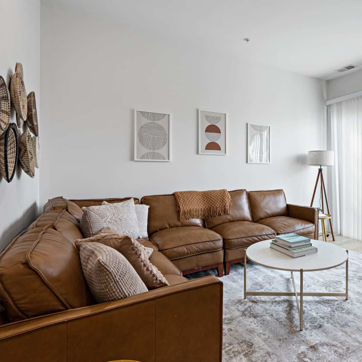 A modern living room featuring a brown leather sectional sofa, a round white coffee table, a standing lamp, and large windows covered with sheer curtains.