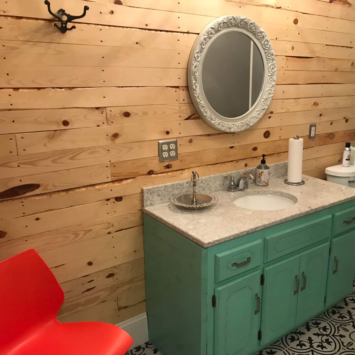 A bathroom featuring a light wood-paneled wall, a turquoise vanity with a countertop sink, a round mirror, a red chair, and a patterned tile floor.