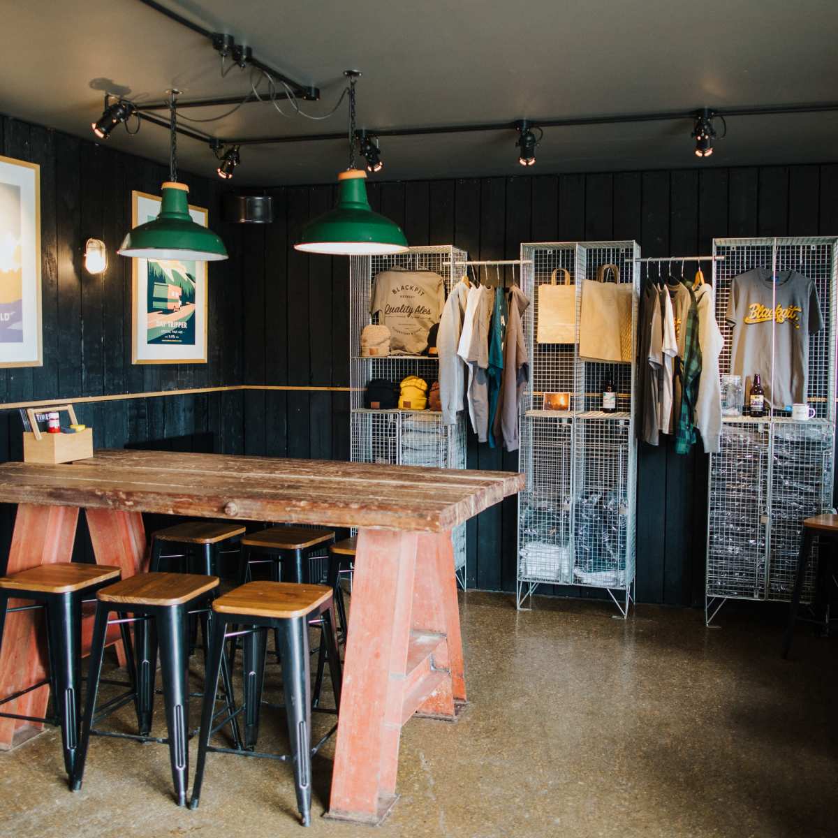 A rustic interior of a shop with a large wooden table, metal stools, and clothing displayed on wire shelving.