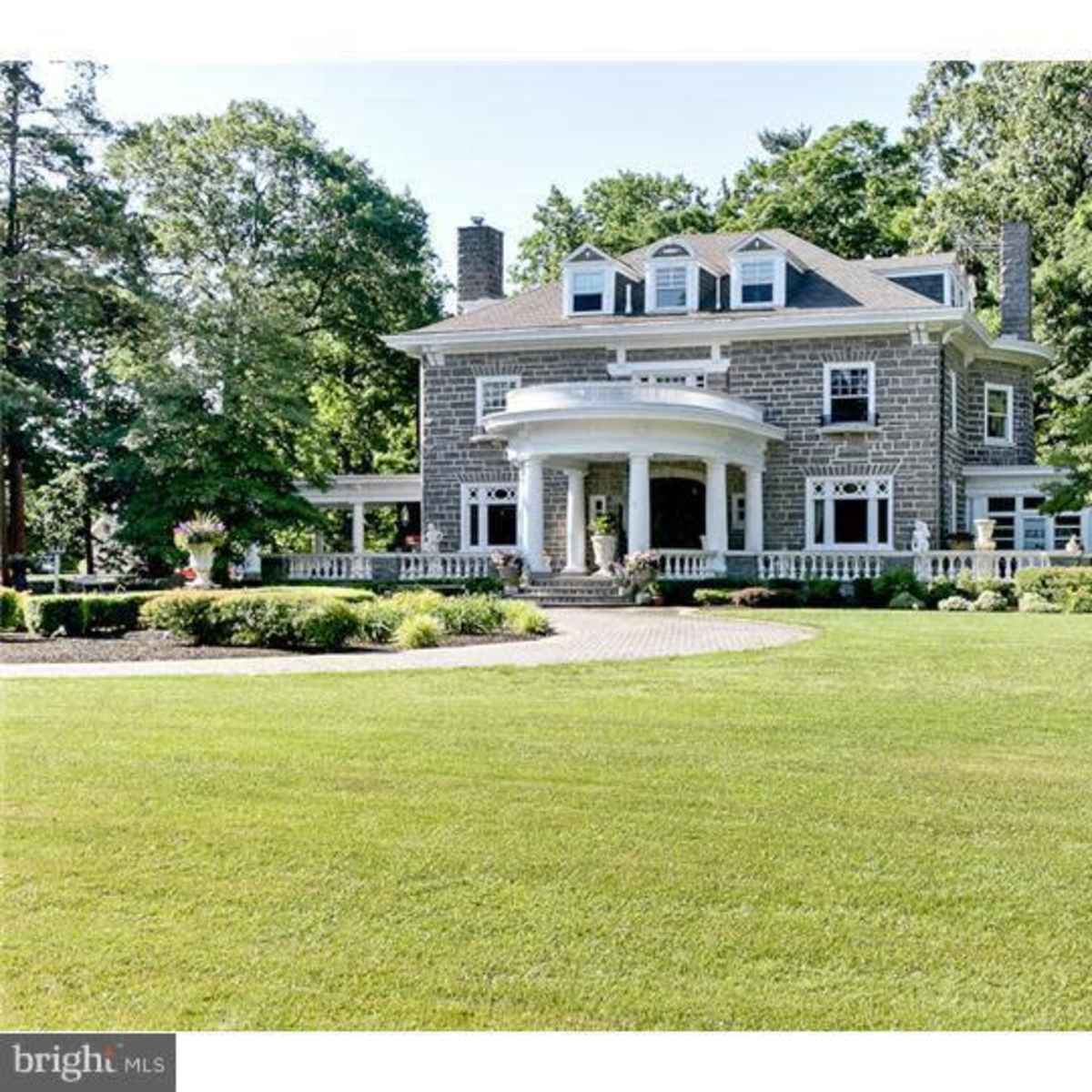 A large, two-story stone house with a circular entrance and a well-maintained lawn surrounded by trees.