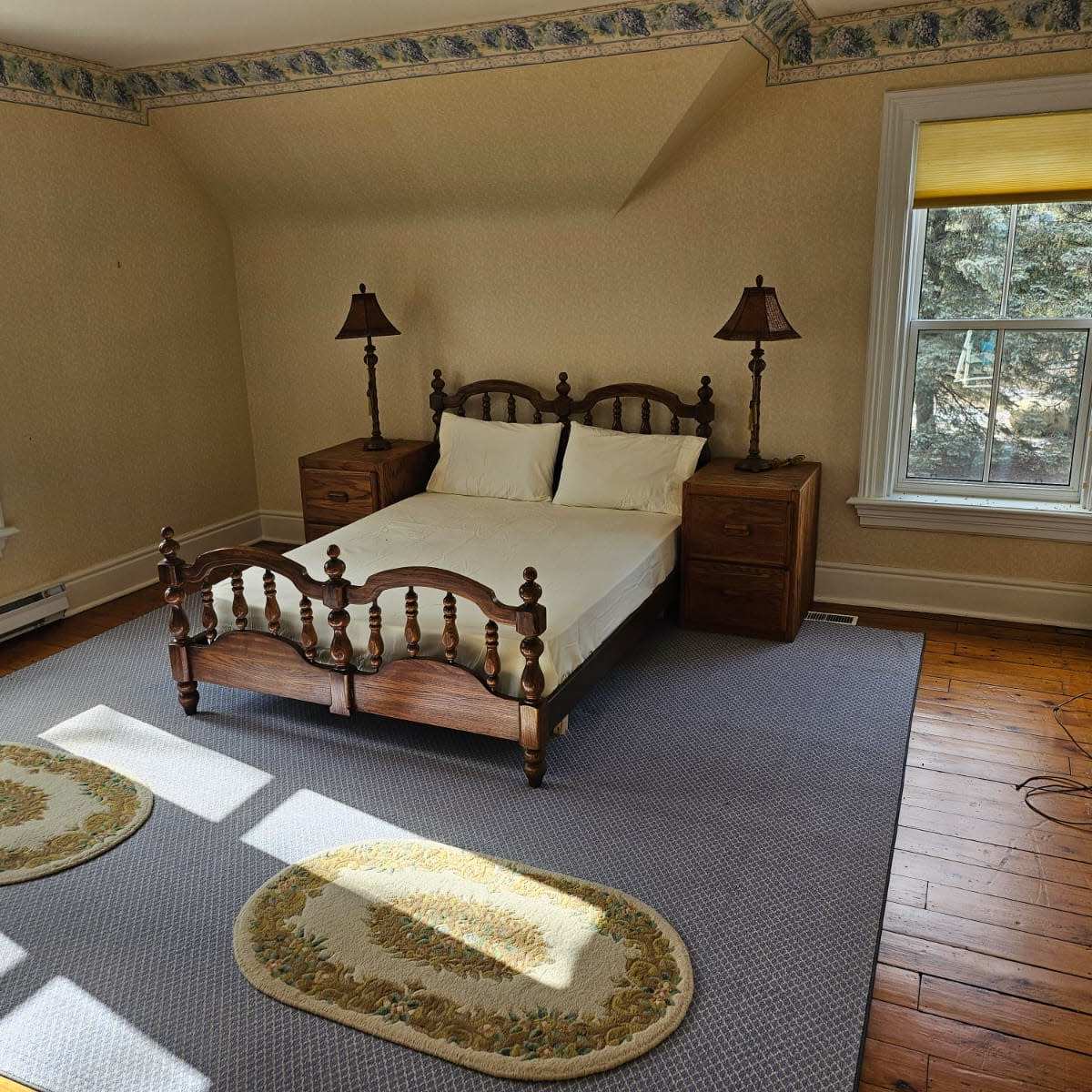 A sparsely furnished bedroom with a wooden bed frame, two lamps on nightstands, and two windows allowing natural light to enter.