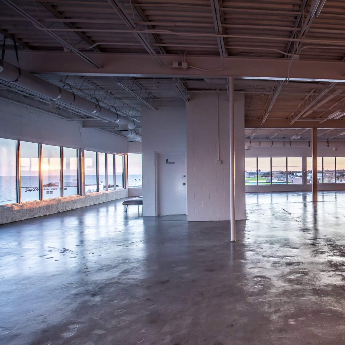 An empty, spacious interior with large windows displaying a view of a waterfront at sunset.