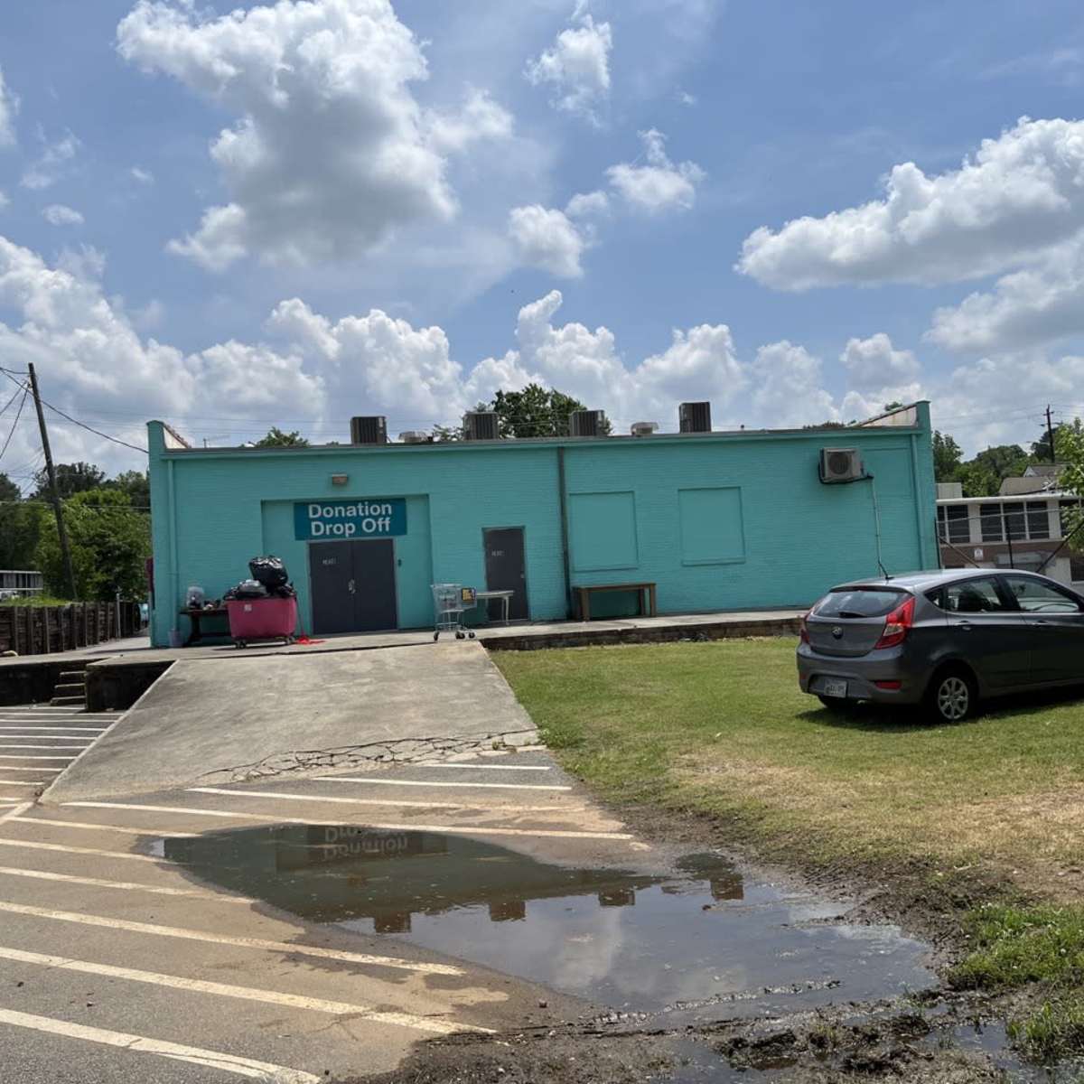 A turquoise building labeled "Donation Drop Off" stands next to a parking area, with a car parked nearby and a puddle on the grass in front.
