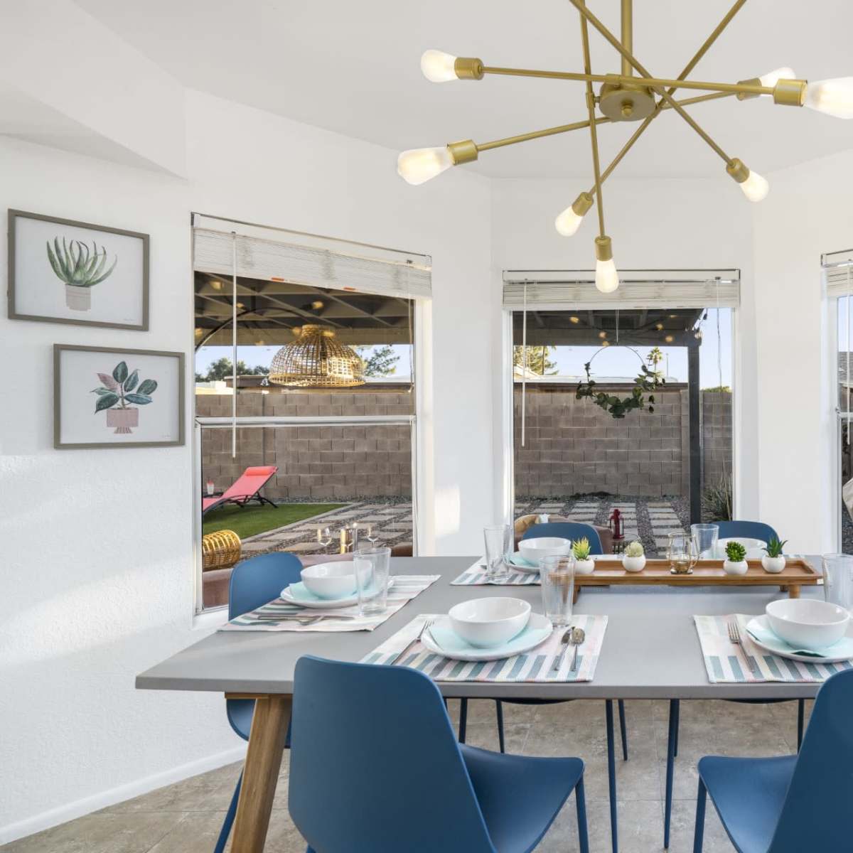 A modern dining area features a table set for six, brightened by a unique light fixture and surrounded by large windows that overlook a patio.