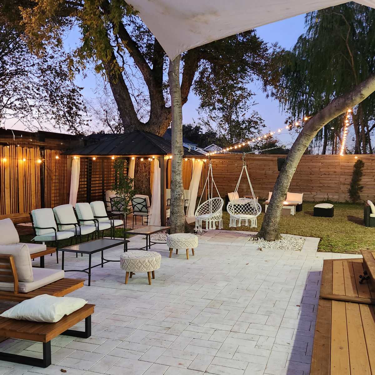 A stylish outdoor lounge area features seating arrangements, decorative lighting, and a wooden deck surrounded by trees.