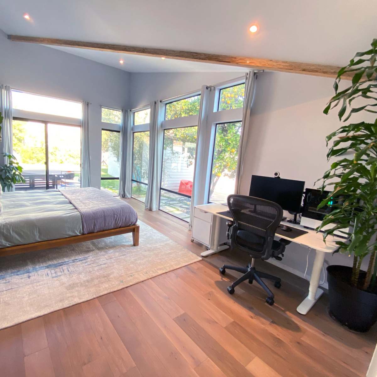 A spacious, well-lit bedroom featuring a bed, a desk with a computer, and large windows overlooking an outdoor area.