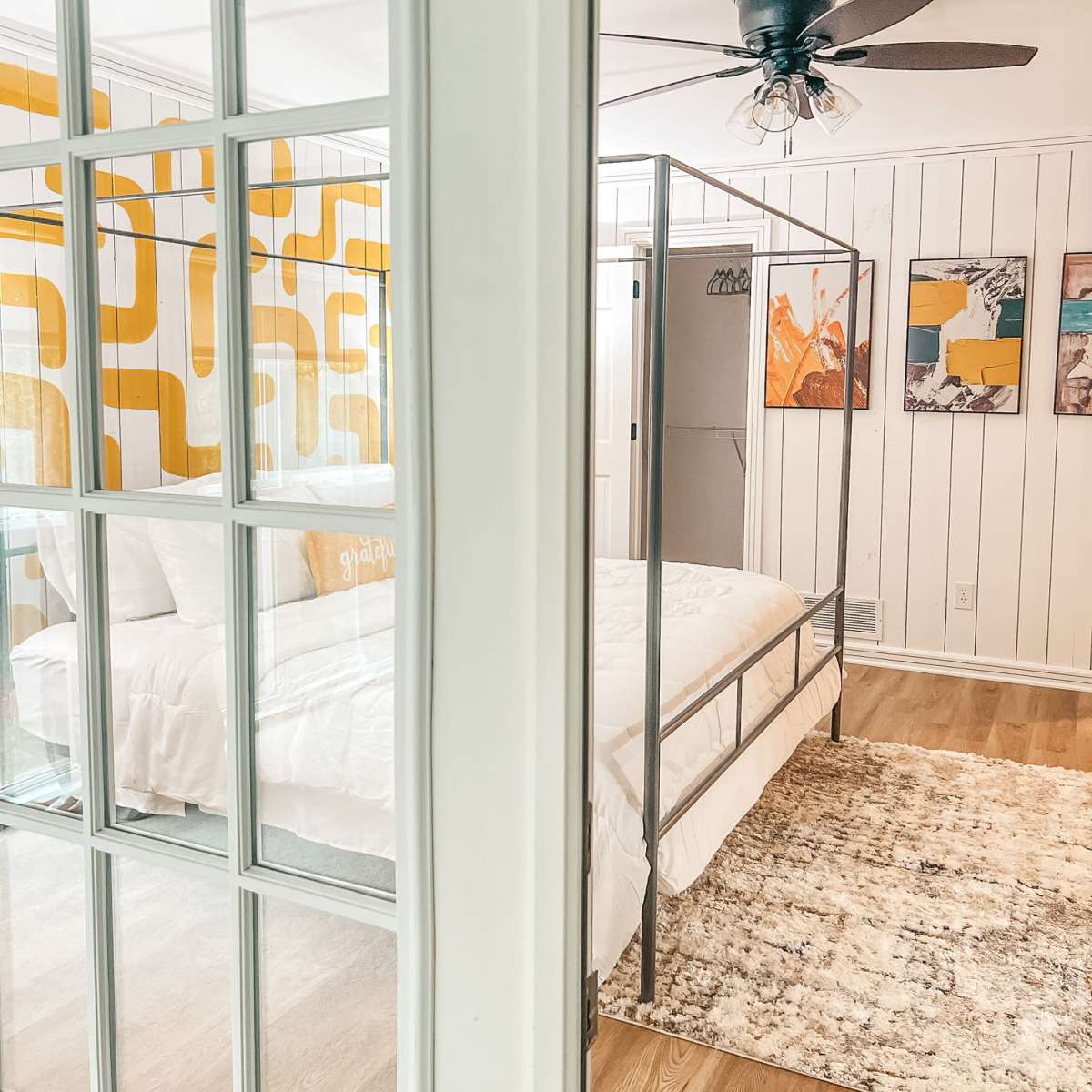 A bright bedroom features a four-poster bed with a white comforter, an accent wall with yellow retro patterns, and framed artwork.