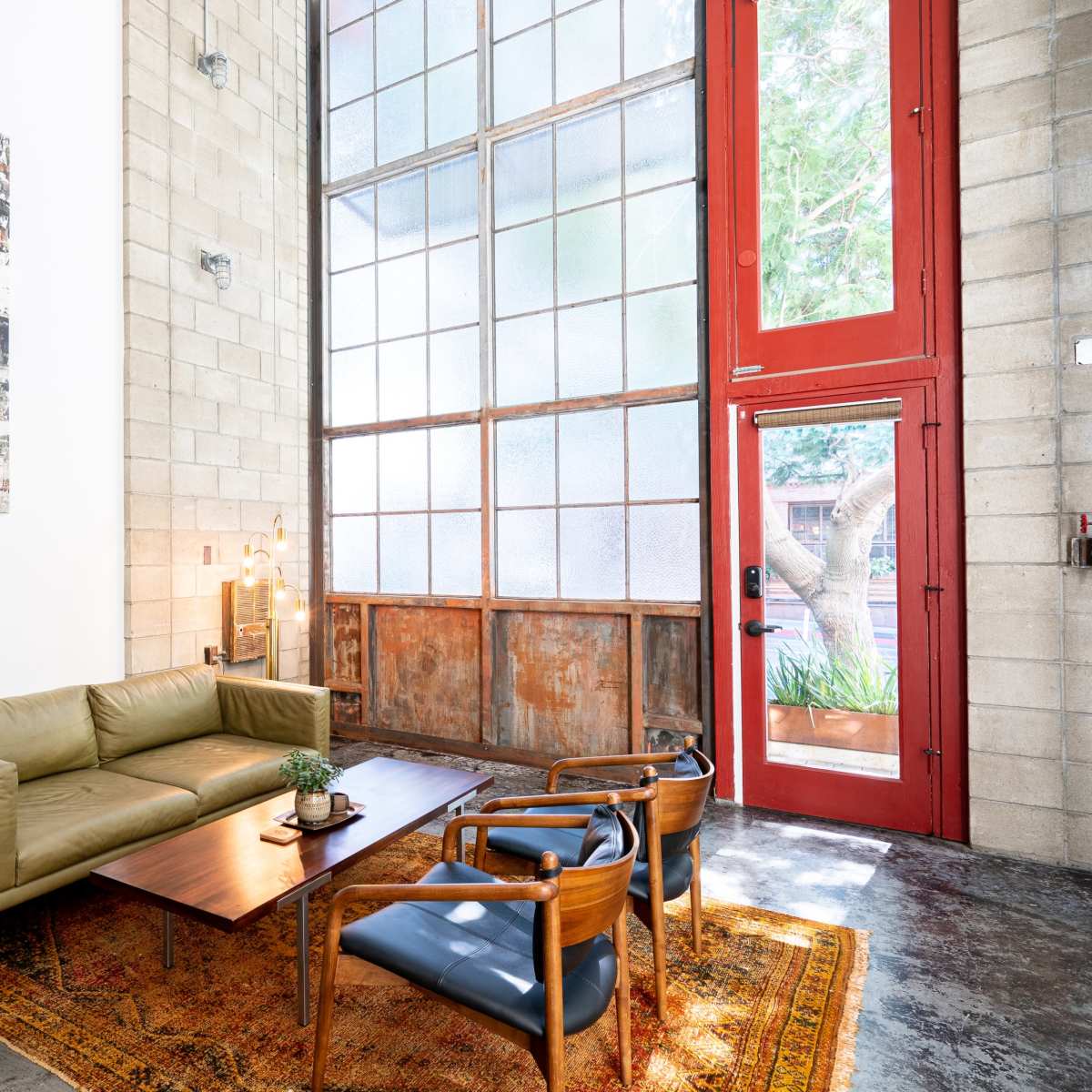 A modern living area with a green sofa, two wooden chairs, a coffee table, and large windows, all set against a backdrop of textured concrete walls.