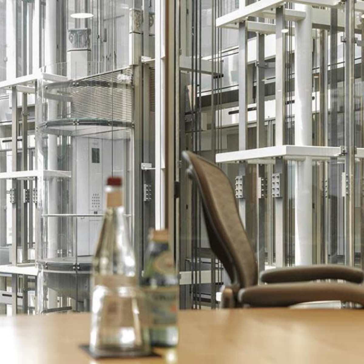 A conference room with a table featuring several glass bottles and a view of an elevator shaft and meeting area through large glass windows.