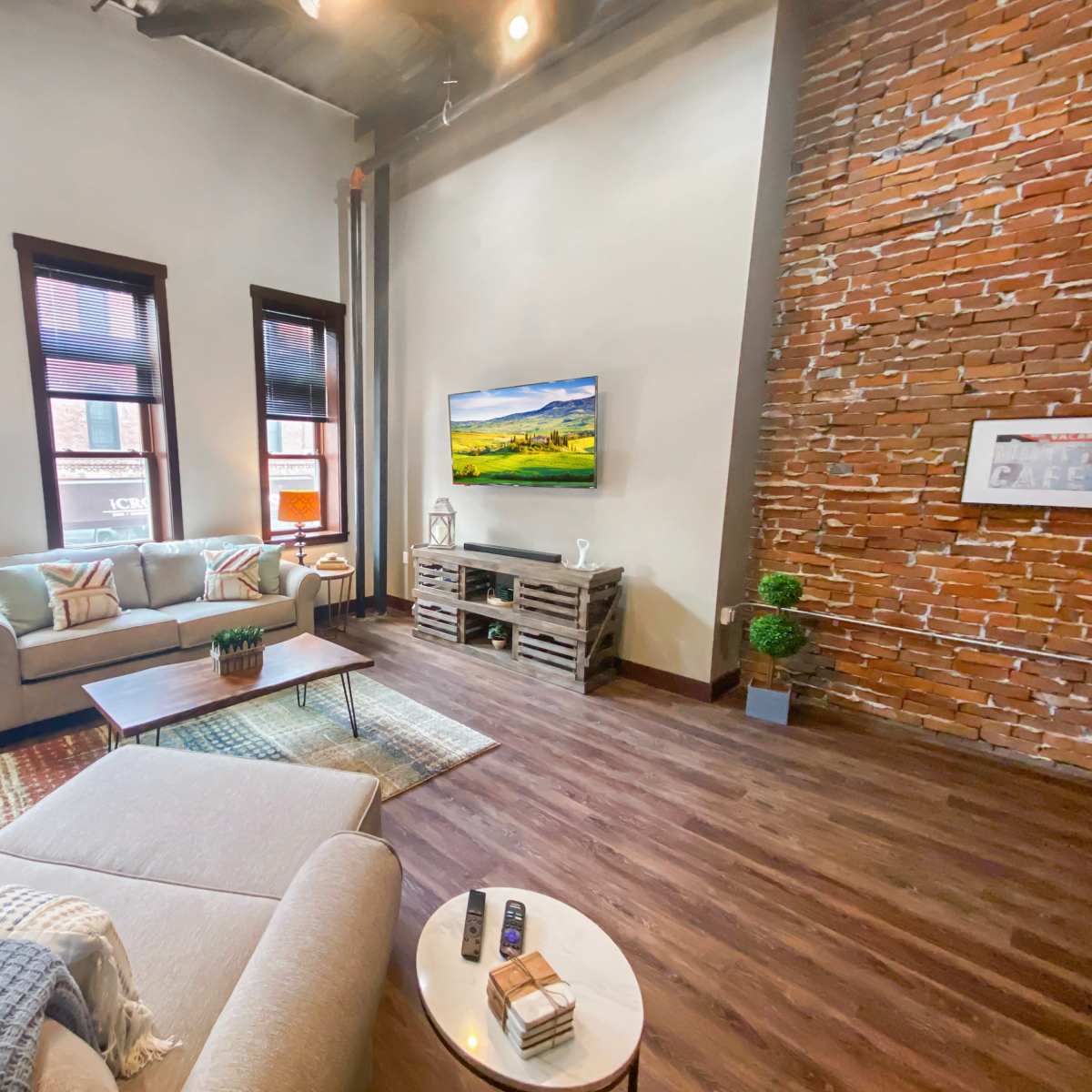 A modern living room featuring a beige sofa, a wooden coffee table, a TV mounted on a brick wall, and large windows with natural light.
