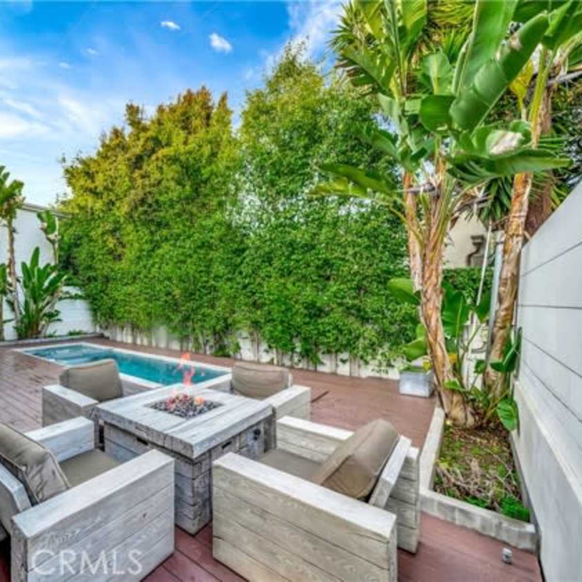 A modern patio area featuring a fire pit surrounded by seating, with a pool and lush greenery in the background.