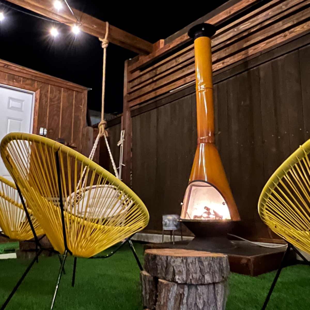 A cozy outdoor space features two yellow chairs facing a chiminea on a wooden platform with green artificial grass.