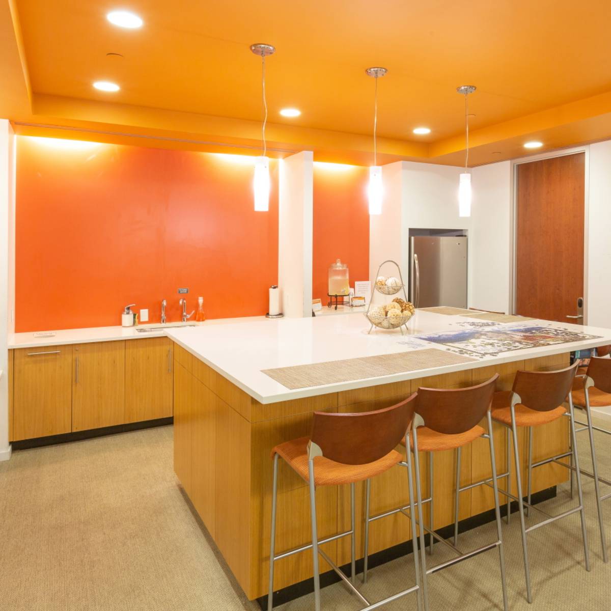 A modern kitchen area with a large island, four bar stools, orange wall accents, and a beverage station.