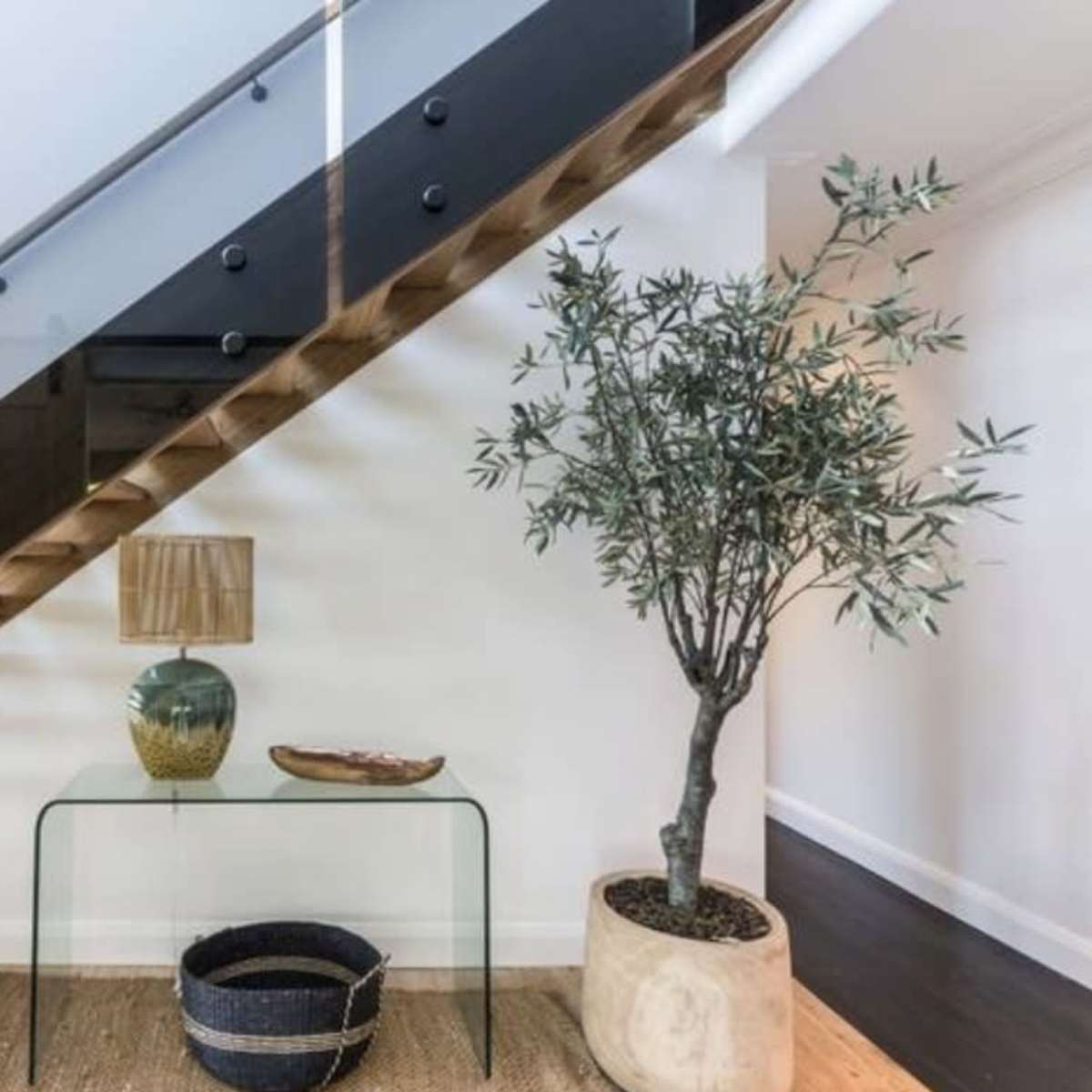 A modern staircase with wooden steps and glass railing features a decorative table with a lamp and a potted plant at its base.
