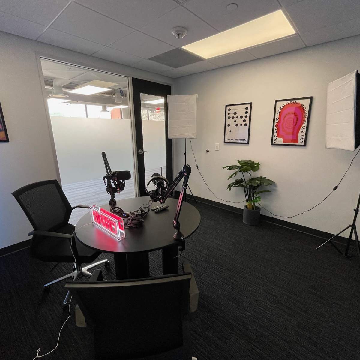 A small recording studio with a round table, two microphones on stands, softbox lights, and artwork on the walls.