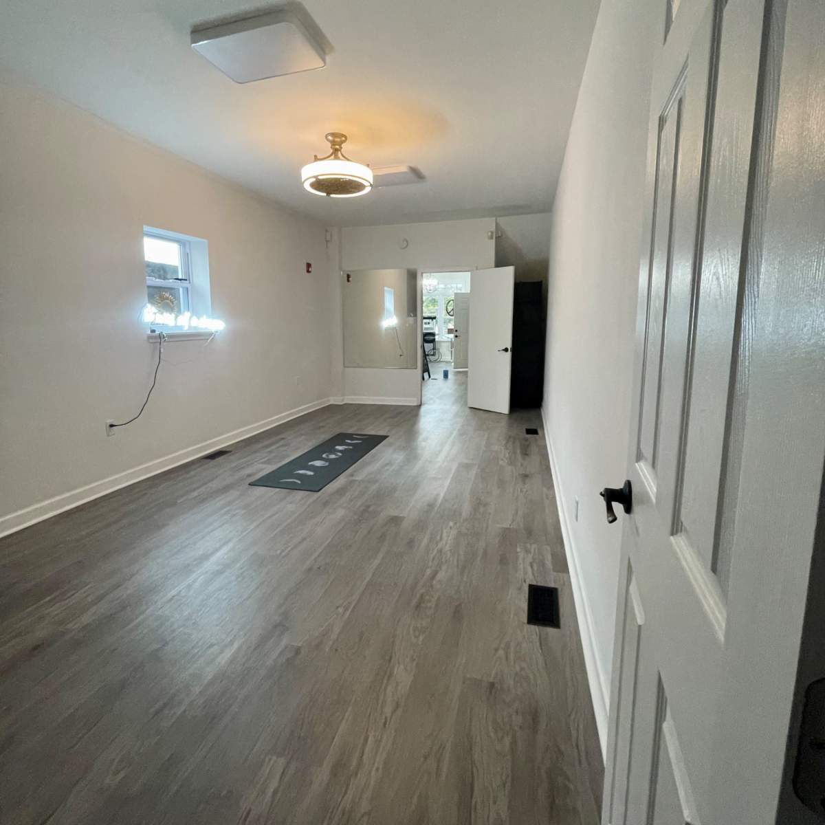 A sparsely furnished room with light-colored walls, a wooden floor, a ceiling light, and a doorway leading to another space.