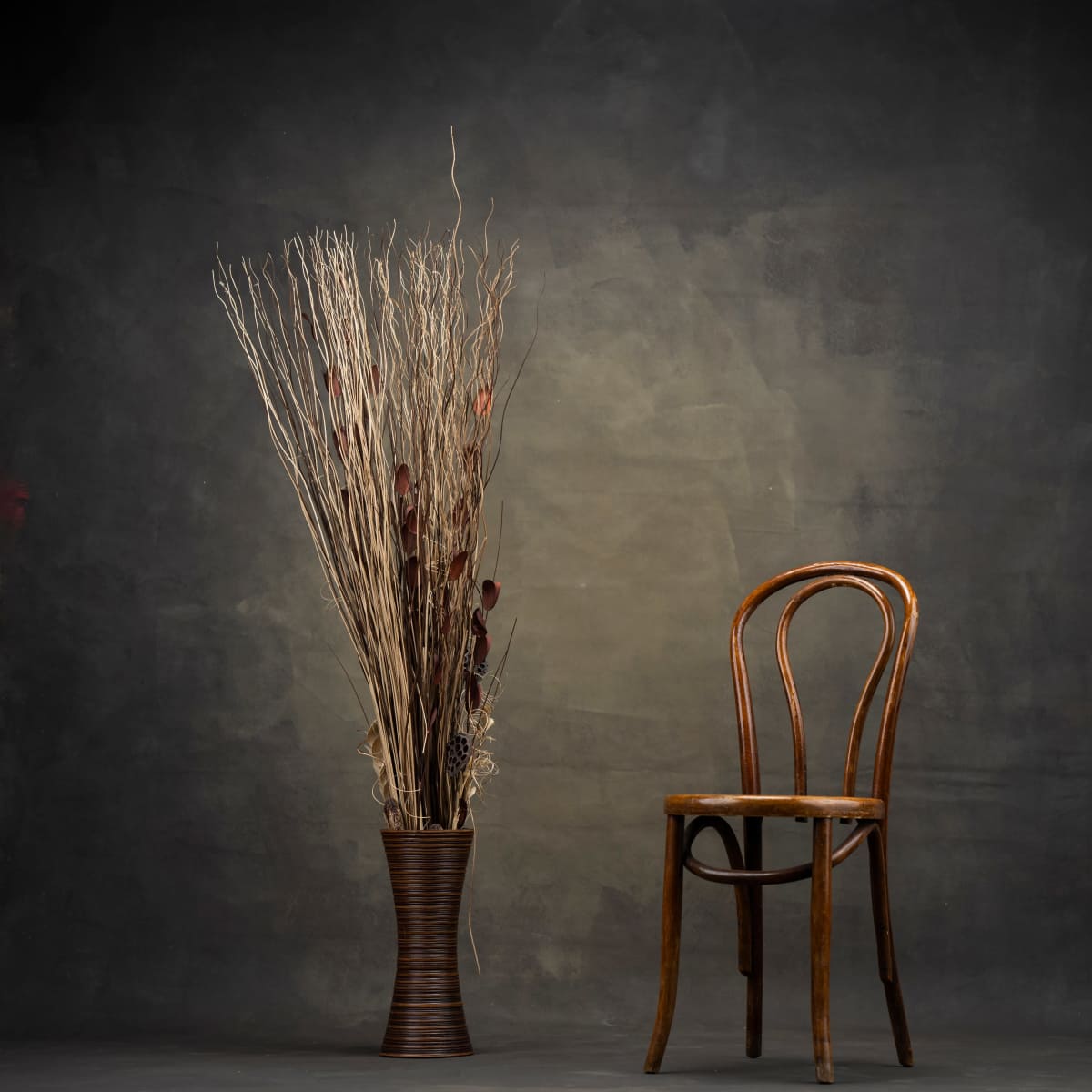 A wooden chair stands beside a tall vase filled with decorative branches against a textured backdrop.