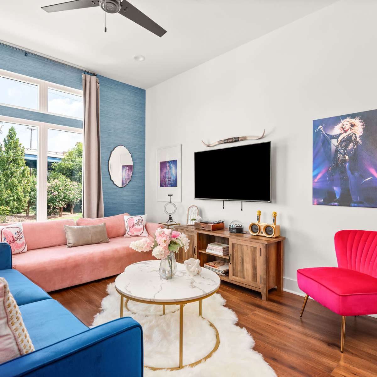 A contemporary living room featuring a pink sofa, a blue couch, a round coffee table, and a TV mounted on the wall, with a large window providing natural light.