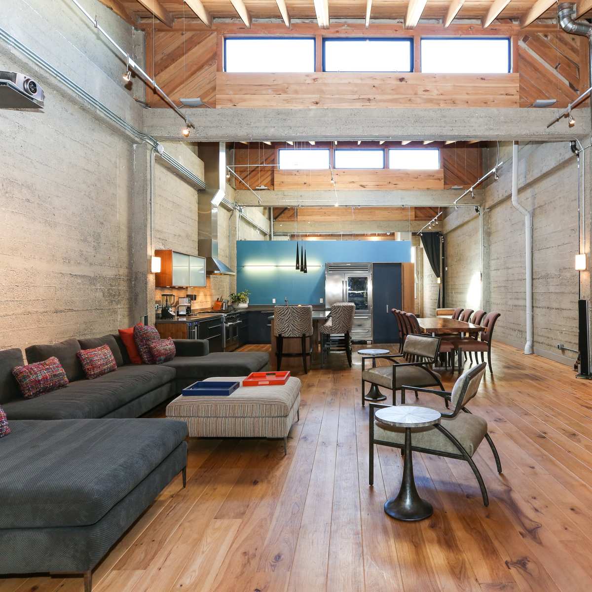 A spacious, modern living area featuring a sectional sofa, a dining table, and a kitchen at the far end, with wooden beams and concrete walls.