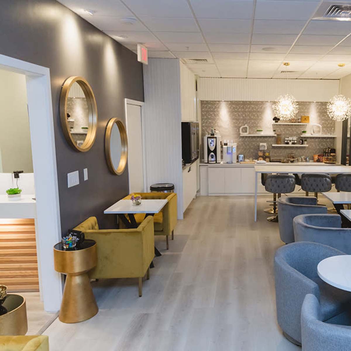 A modern lounge area with gray walls, circular mirrors, a kitchenette, and various seating arrangements including chairs and tables.