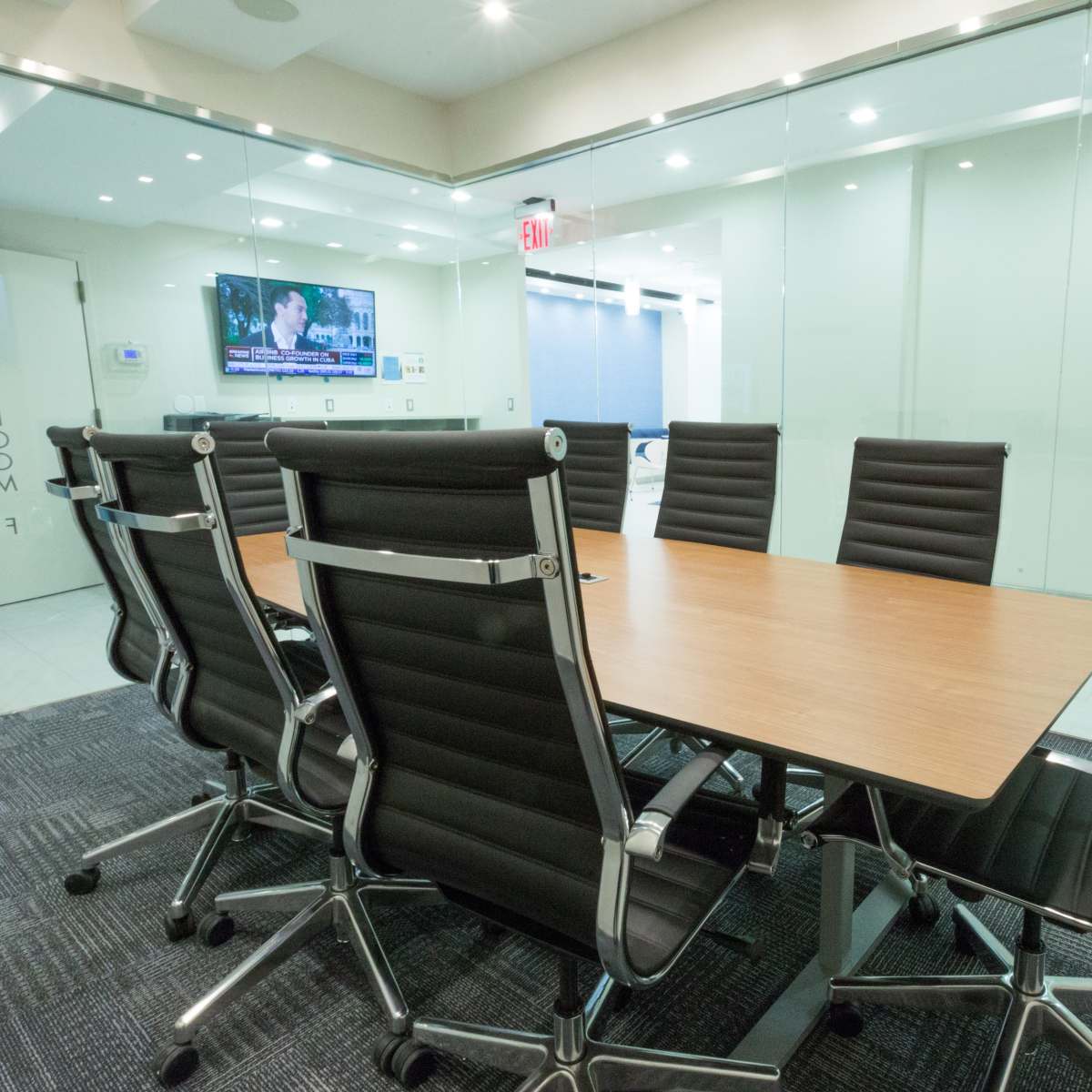 A modern conference room features a large wooden table surrounded by black ergonomic chairs, with a TV mounted on the wall displaying news.