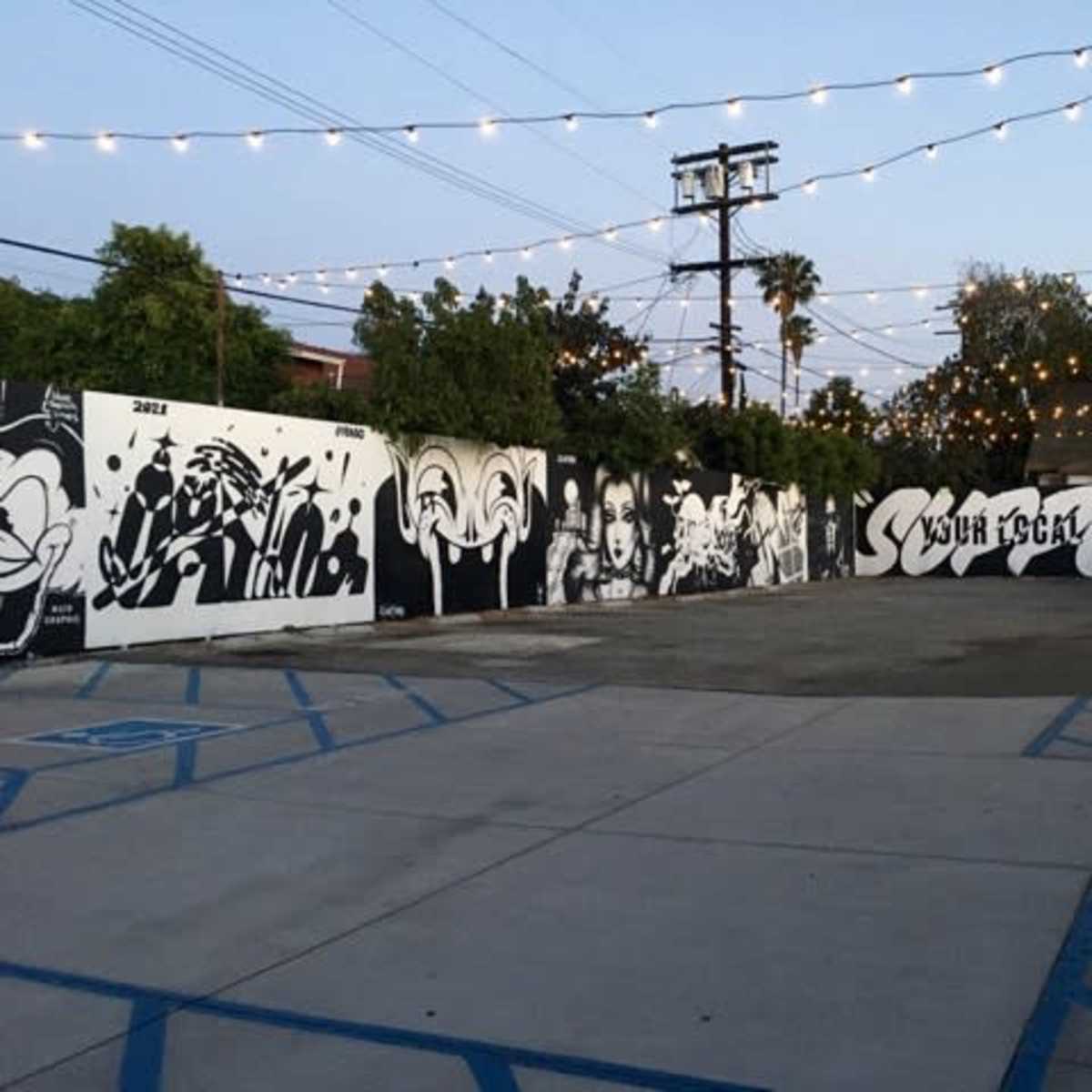 An outdoor mural-covered area with string lights above, featuring several large black and white artworks on the walls.